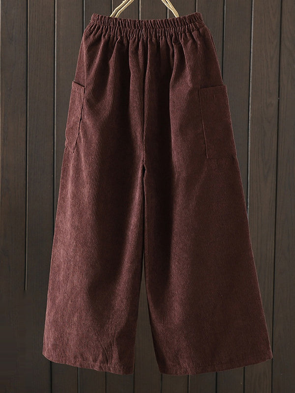 A pair of dark brown corduroy wide-leg pants with an elastic waistband and side pockets, hanging against a dark wood plank background.