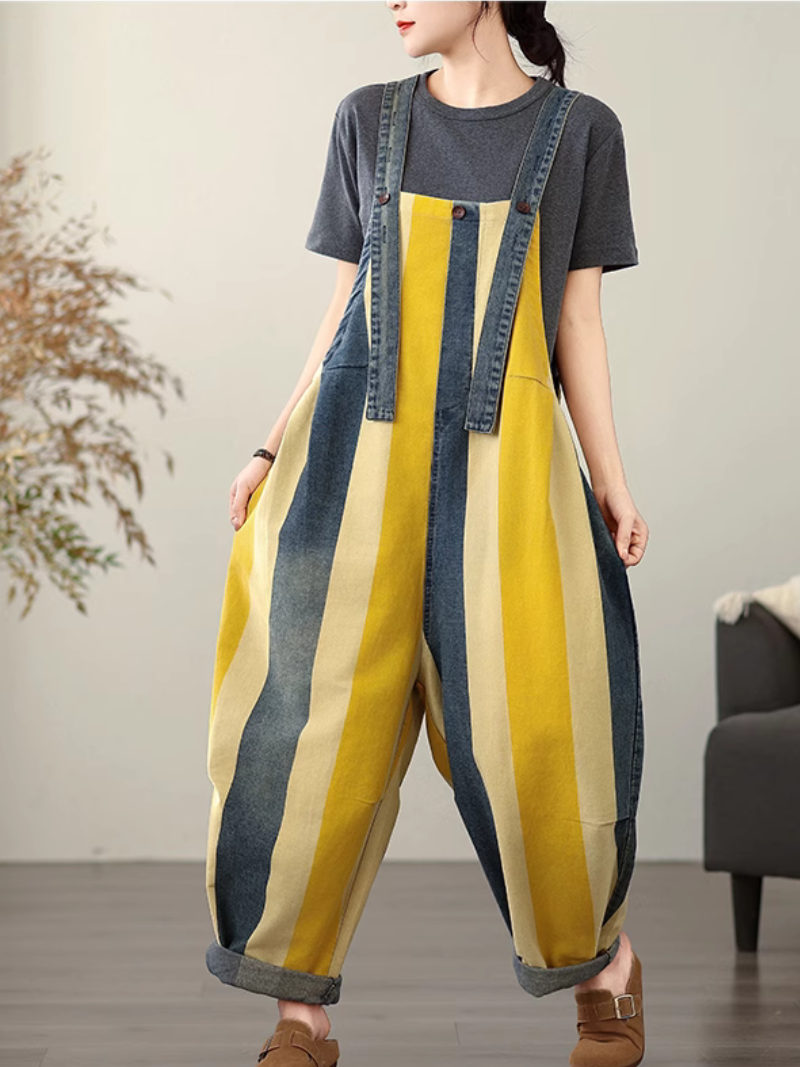 A woman wearing a striped colorful dungarees over a grey shirt with denim details, standing indoors.