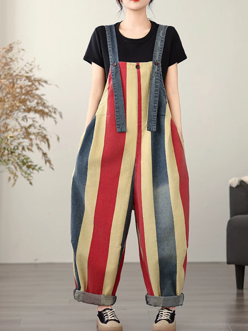 A woman wearing a striped colorful dungarees over a grey shirt with denim details, standing indoors.