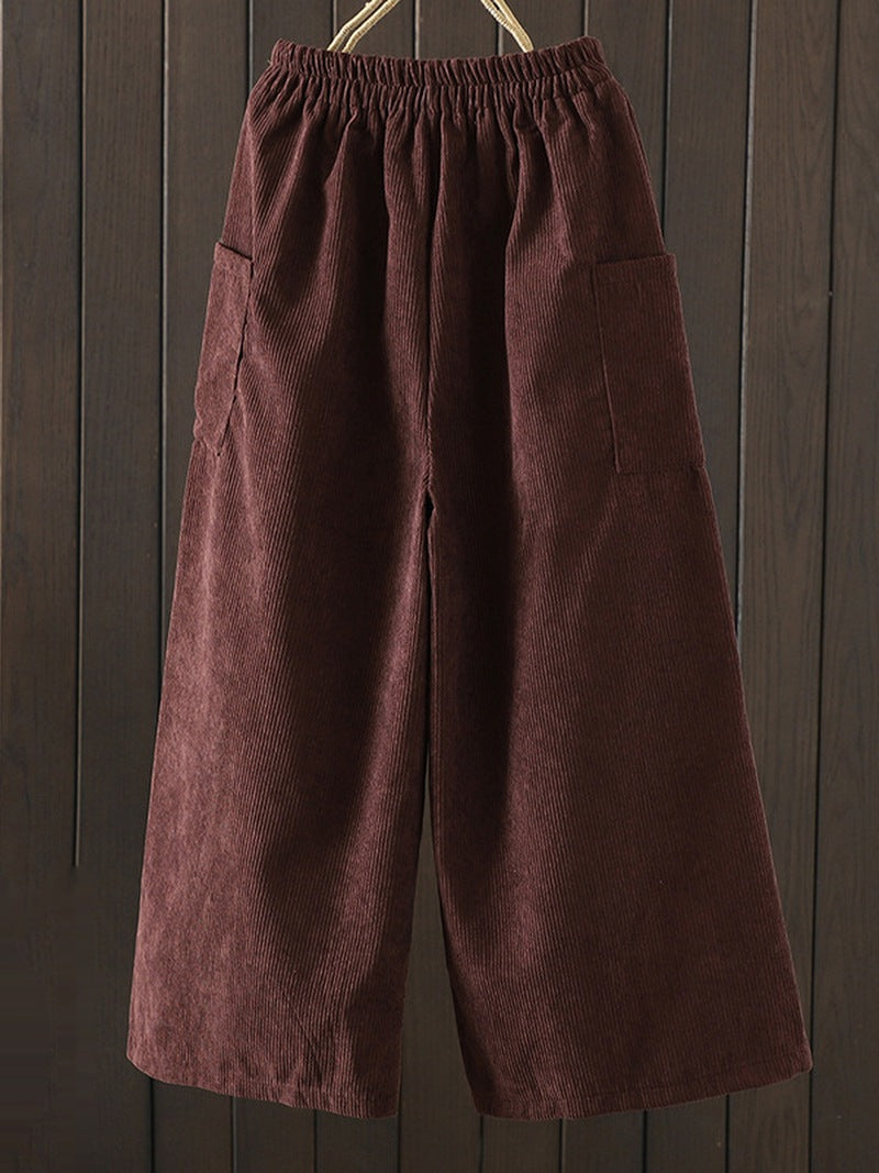 A pair of dark brown corduroy wide-leg pants with an elastic waistband and side pockets, hanging against a dark wood plank background.